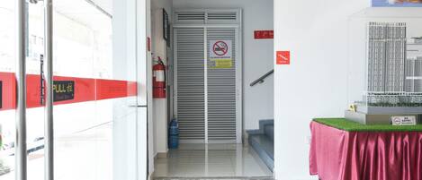 Entrada de un hotel con extintor, un cartel de prohibido fumar y puerta de cristal que da a una escalera.