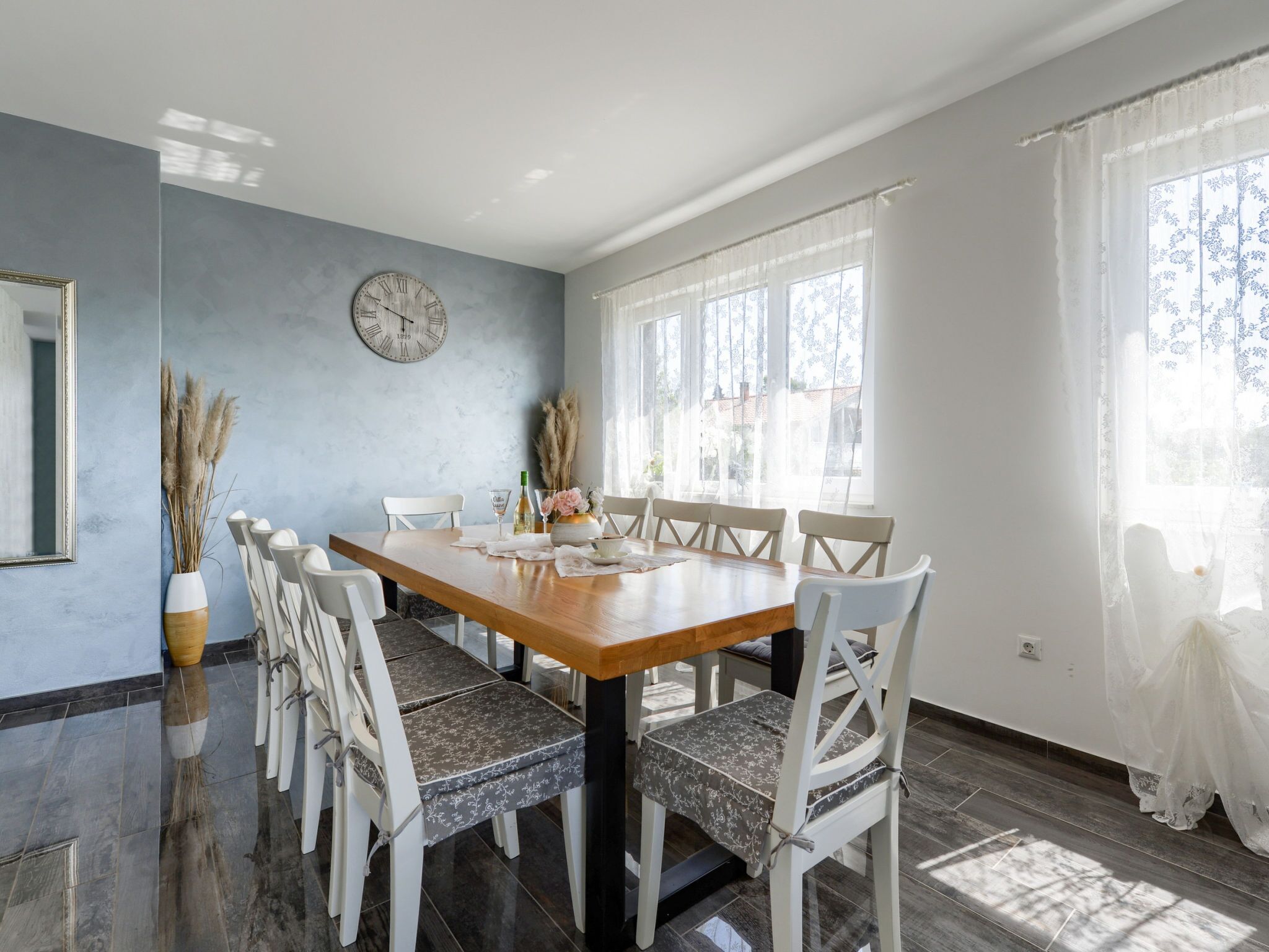 A dining room with a wooden table, white chairs, a clock on the wall, and large windows with sheer curtains.