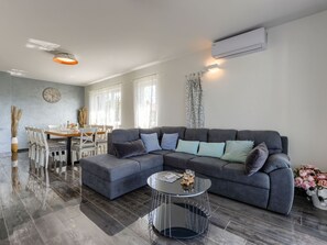Ein modernes Wohnzimmer mit einer Eckcouch, einem Essbereich mit Tisch und Stühlen sowie einem großen Fenster, das für viel Tageslicht sorgt.