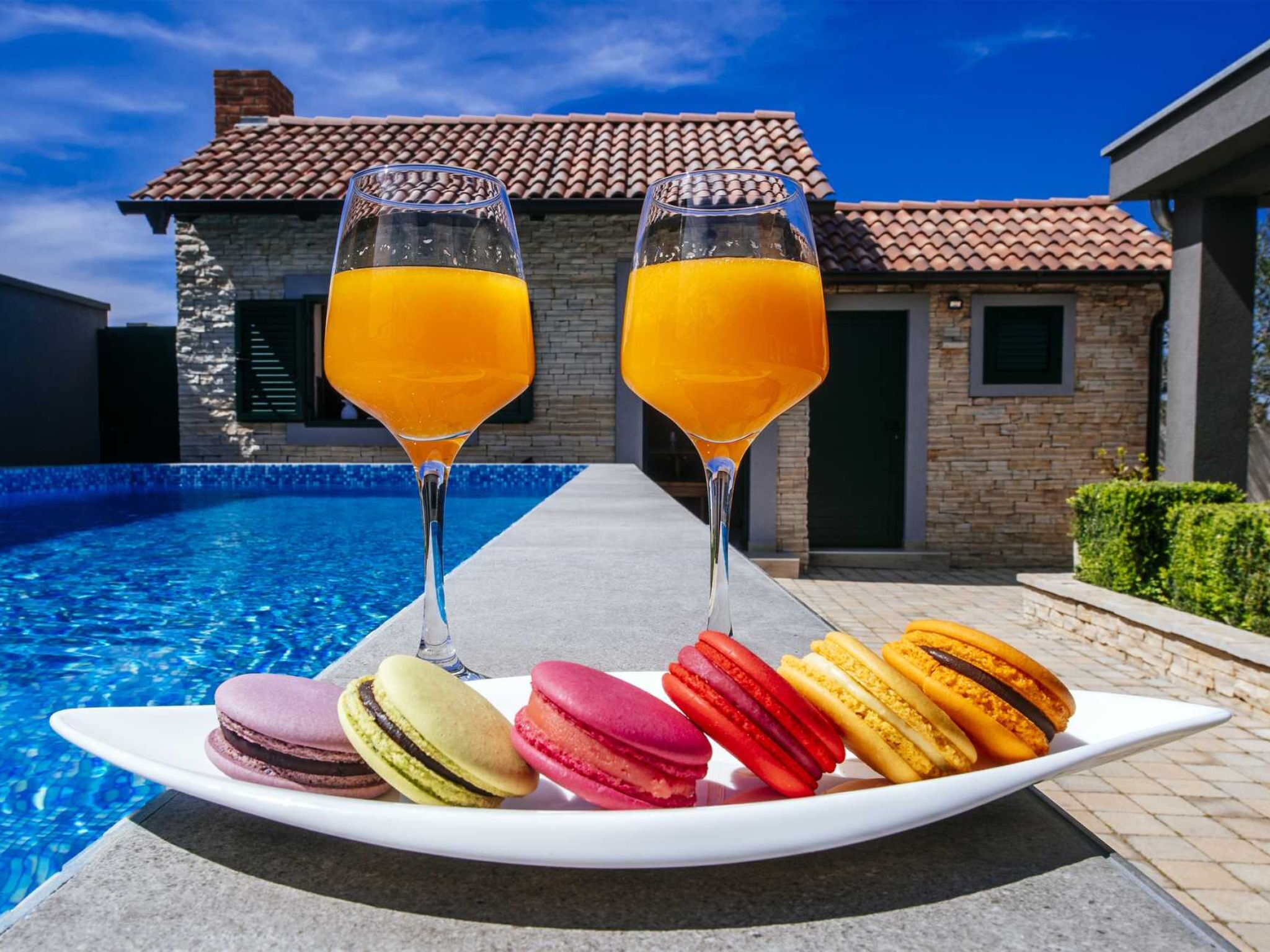 A poolside setting with two glasses of orange juice, a plate of colorful macarons, and a building with a tiled roof in the background.