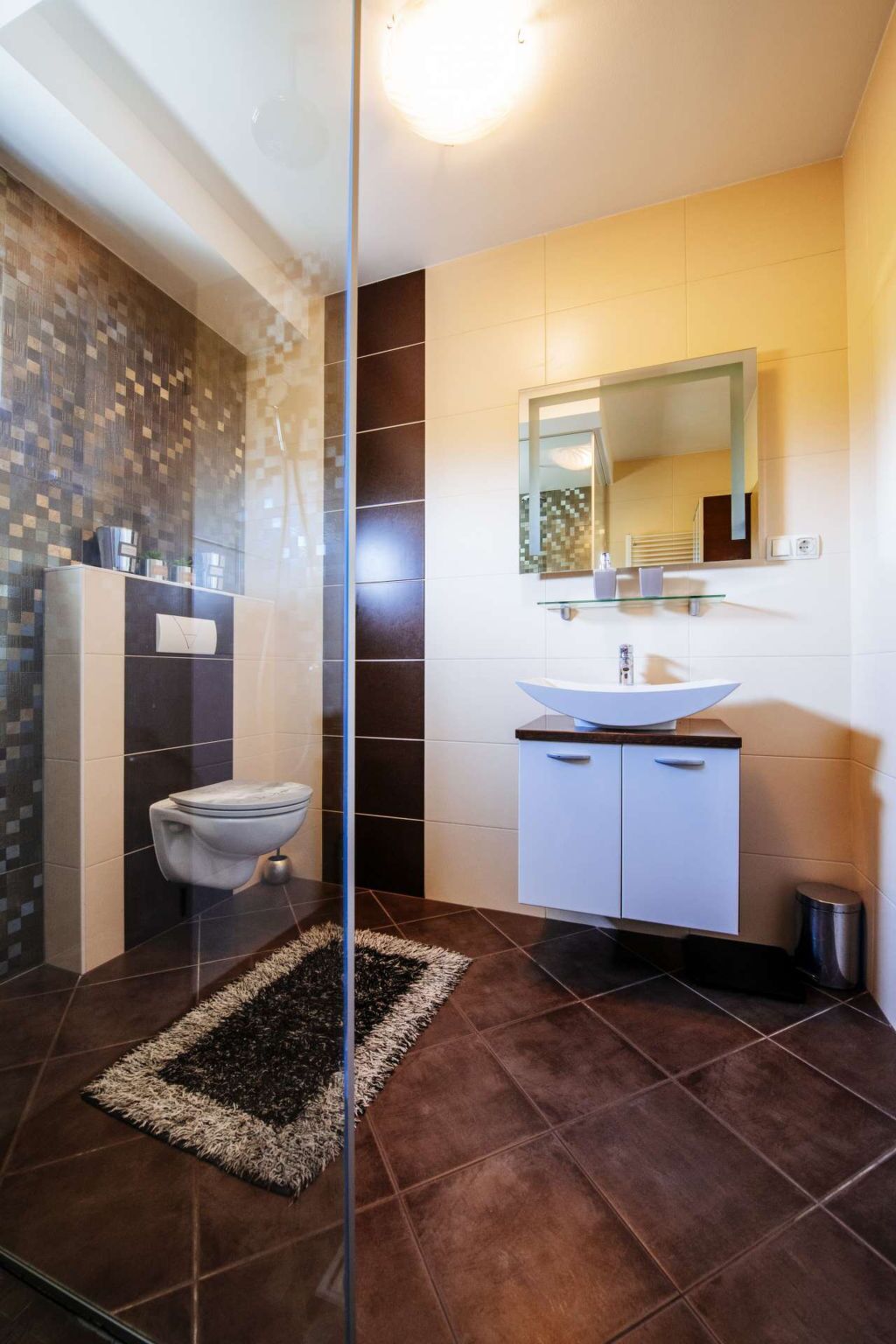 Un baño moderno con cabina de ducha de vidrio, inodoro, lavamanos con espejo y un cubo de basura pequeño.