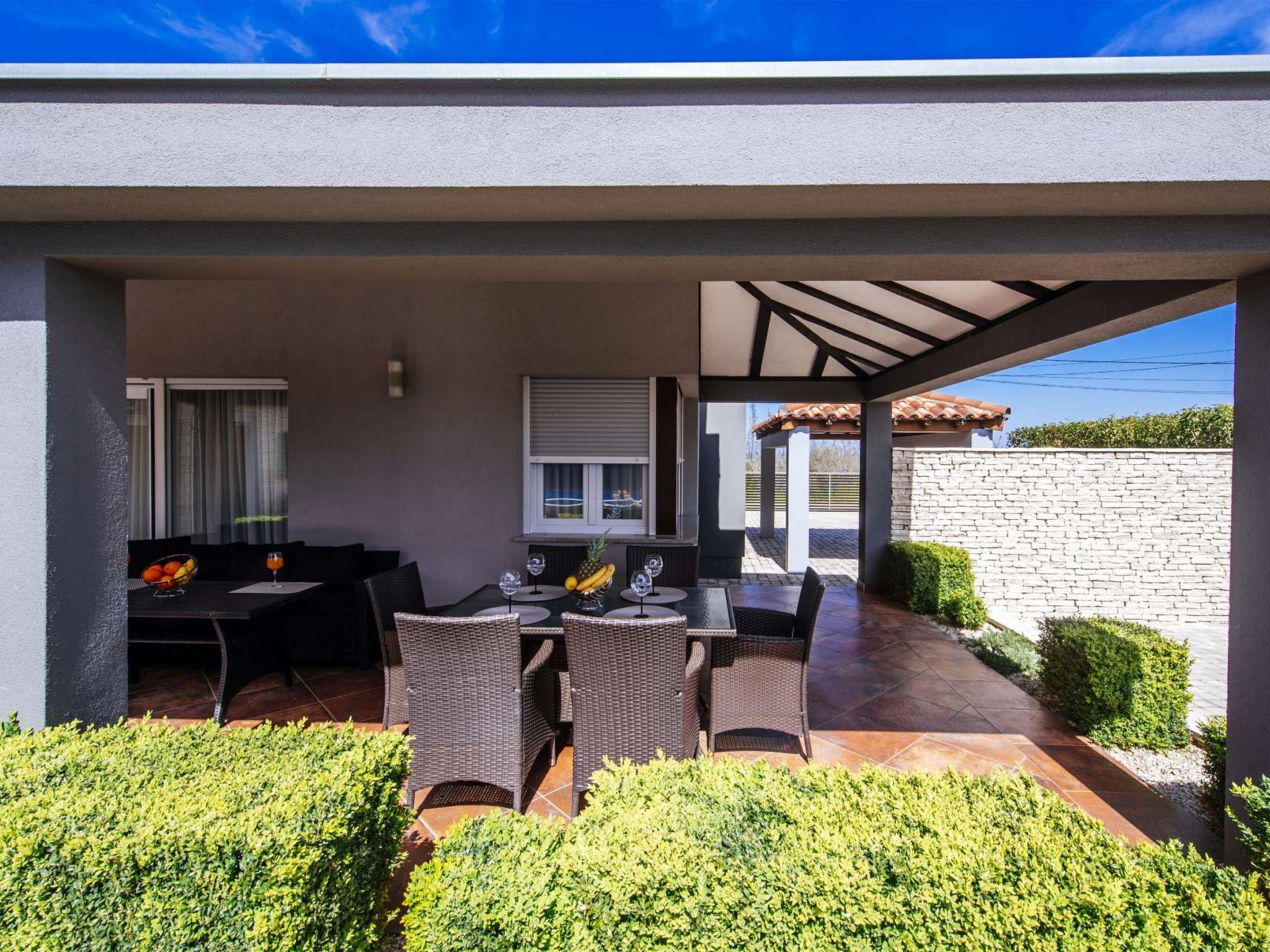 A covered outdoor dining area with a table and chairs, a window, and a stone wall in the background.