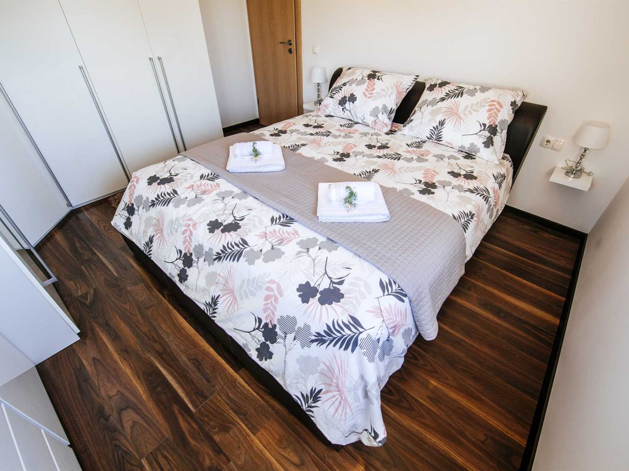 A bed with floral bedding, a bedside table with a lamp, and a wooden floor.