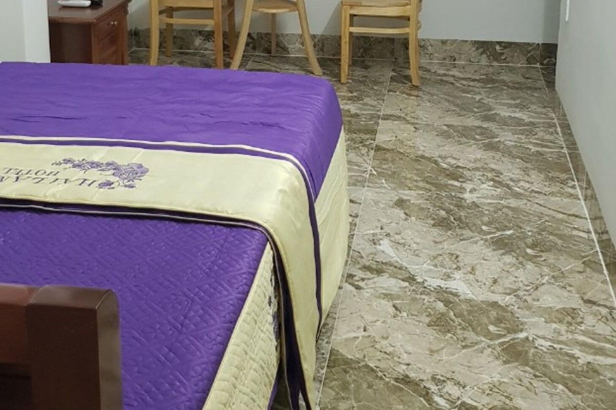 Una stanza con un letto, due poltrone, un tavolino, una finestra con tende viola e un punto luce a parete.