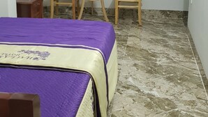 Una habitación con una cama, dos sillas, una mesita, una ventana con cortinas moradas y una lámpara fijada a la pared.