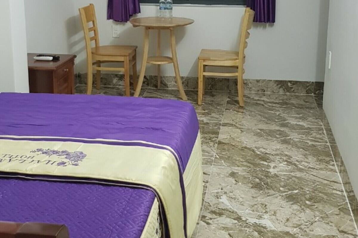 A room with a bed, two chairs, a small table, a window with purple curtains, and a wall-mounted light fixture.