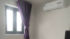 Una habitación con ventana, cortinas moradas, una silla de madera, una mesita redonda y un aire acondicionado instalado en la pared.