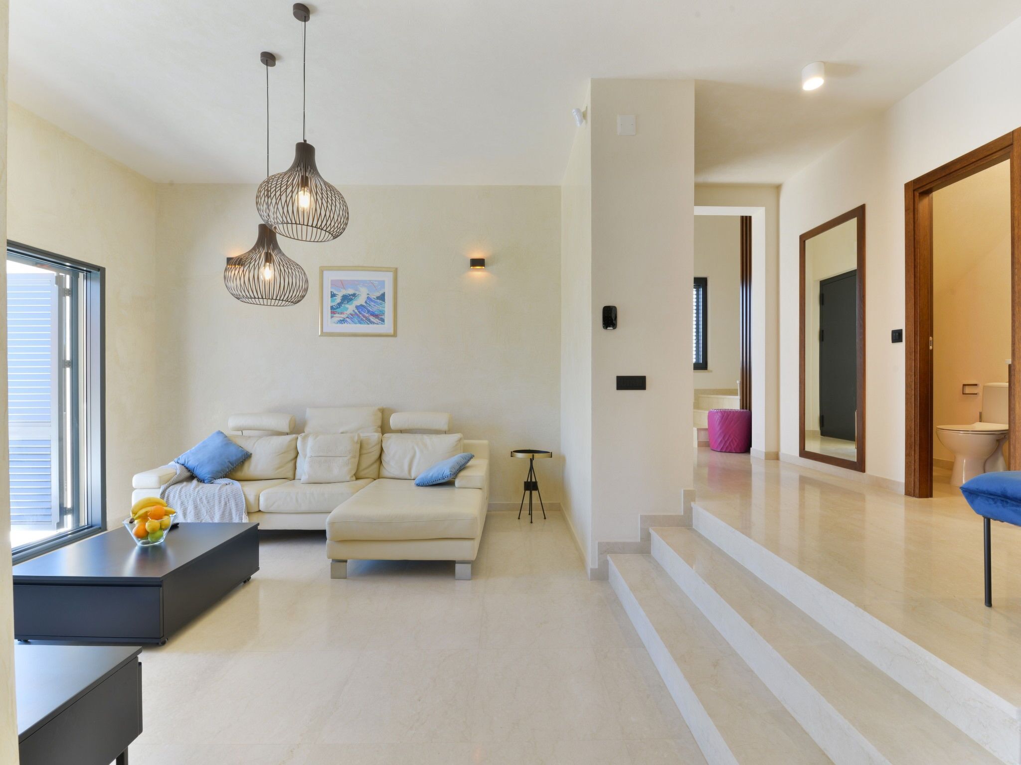 A modern living room with a sofa, a coffee table, and a fruit bowl. There is a staircase leading to another level.