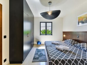 Ein modernes Schlafzimmer mit einem Bett, einem blauen Hocker, einem Fenster mit Blick und einer Deckenleuchte.