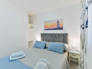 Ein Schlafzimmer mit einem Bett, Kissen, einem Nachttisch, einer Lampe, einem Gemälde eines Leuchtturms und einem Vorhang mit Meeresmotiv.
