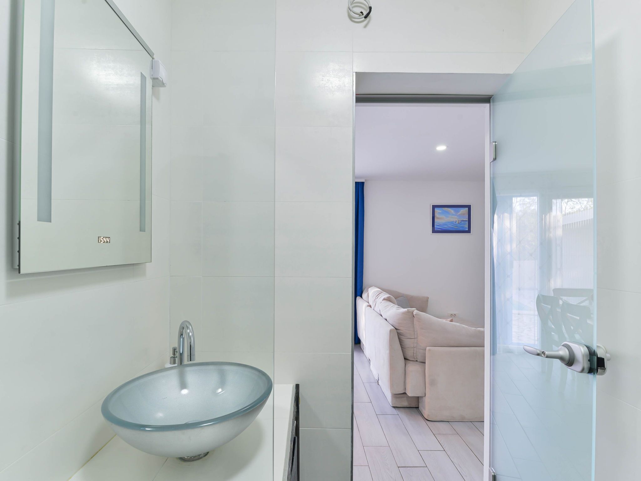Une salle de bain moderne avec un lavabo en verre, un miroir et une porte donnant sur un espace de vie comprenant un canapé et un tableau.