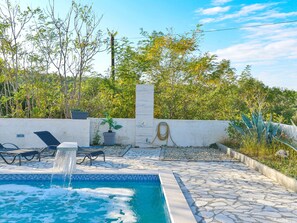 Ein Schwimmbecken mit Wasserspiel, umrahmt von Steinplatten, Liegestühlen und einem Gartenbereich.