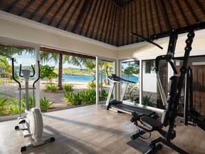 A gym with exercise equipment, large windows, and a view of the beach. - Sudamala Resort, Seraya (Labuan Bajo)