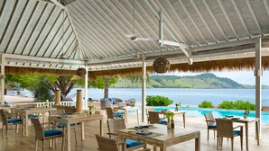 A covered outdoor seating area with tables and chairs, overlooking a pool and a scenic view of a body of water and hills. - Sudamala Resort, Seraya (Labuan Bajo)