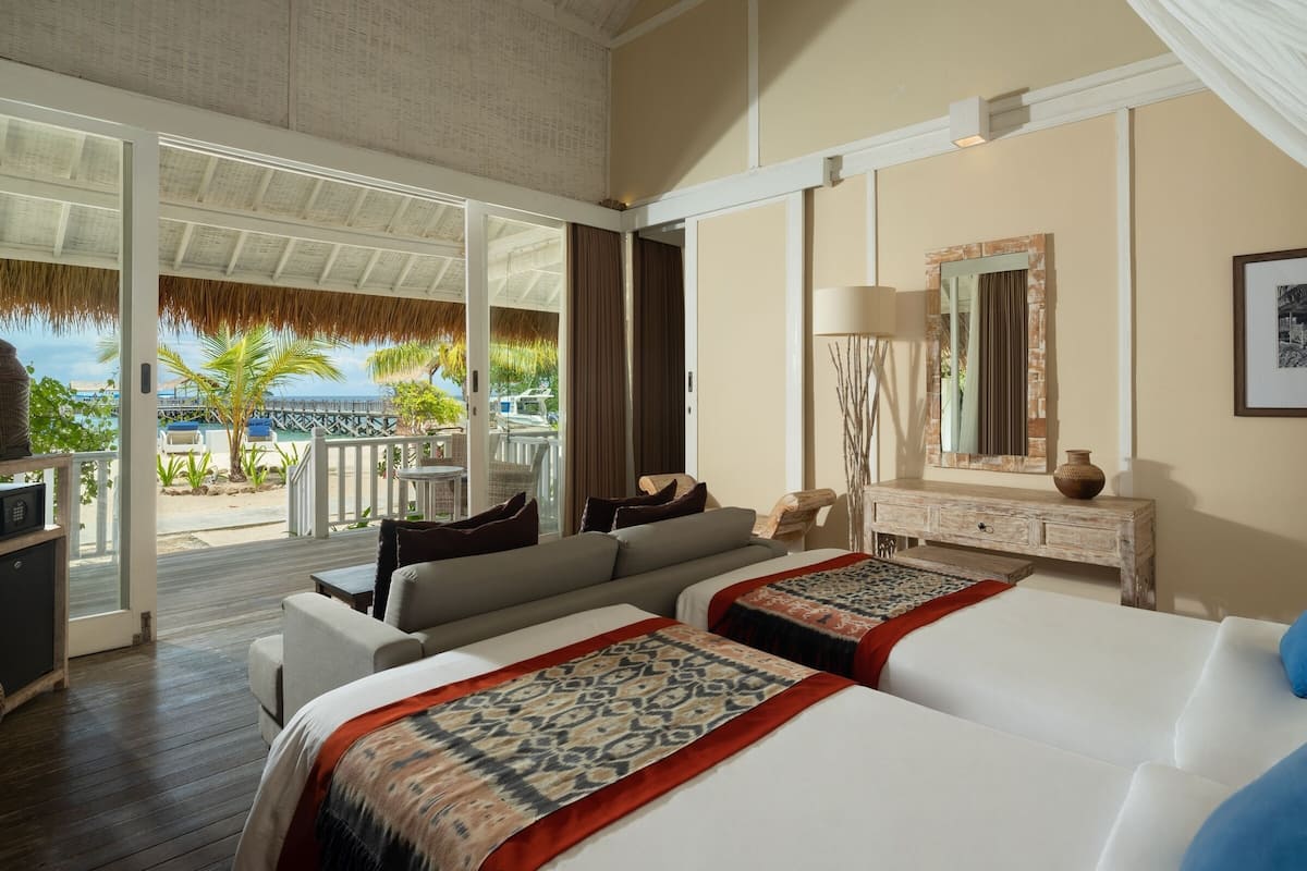 Habitación de hotel con un amplio ventanal corredera que da a un balcón, una cama con un plaid estampado, un aparador de madera y un espejo con marco decorativo.