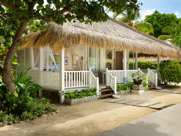 A thatched-roof bungalow with a white porch, surrounded by tropical vegetation.