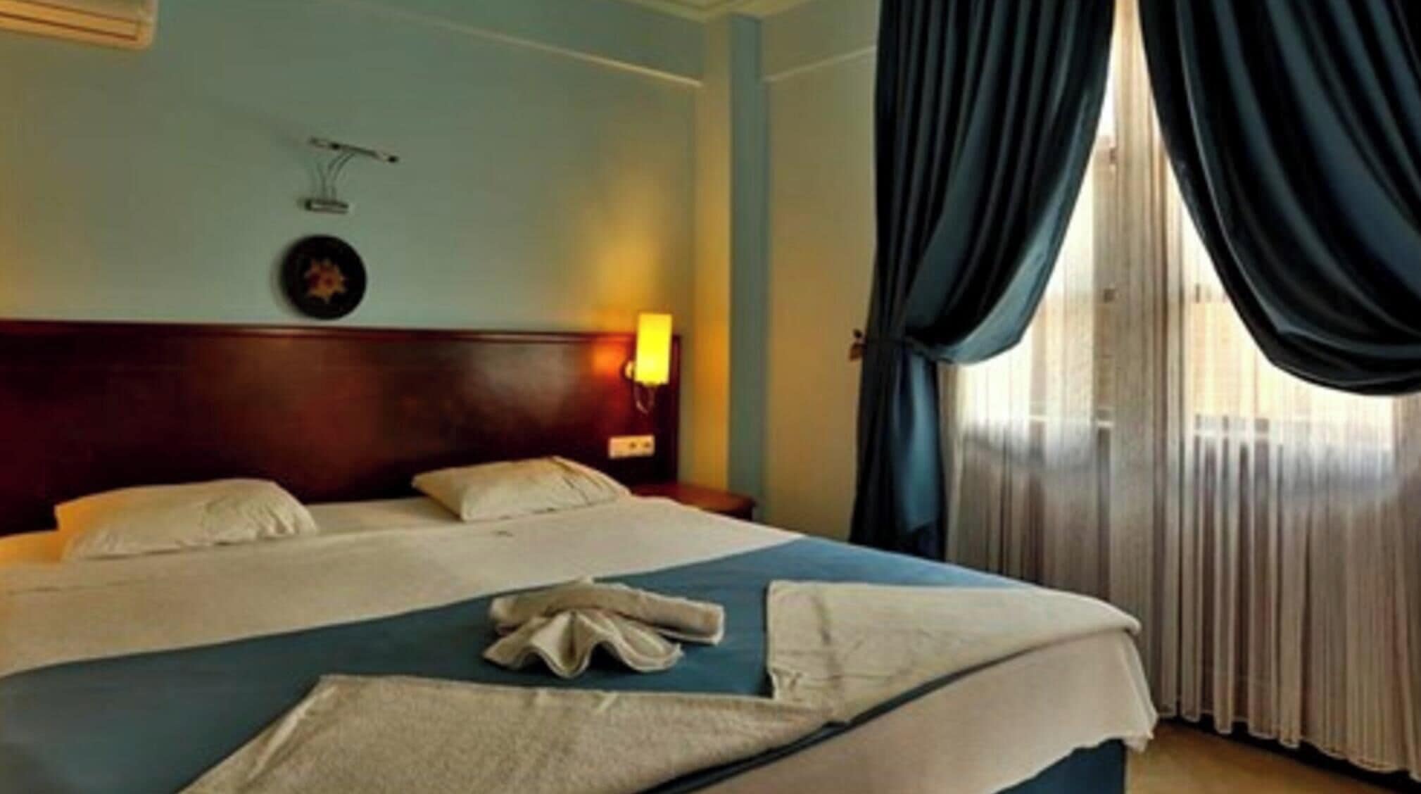 A hotel room with a large bed, a bedside lamp, a clock on the wall, and a window with curtains.