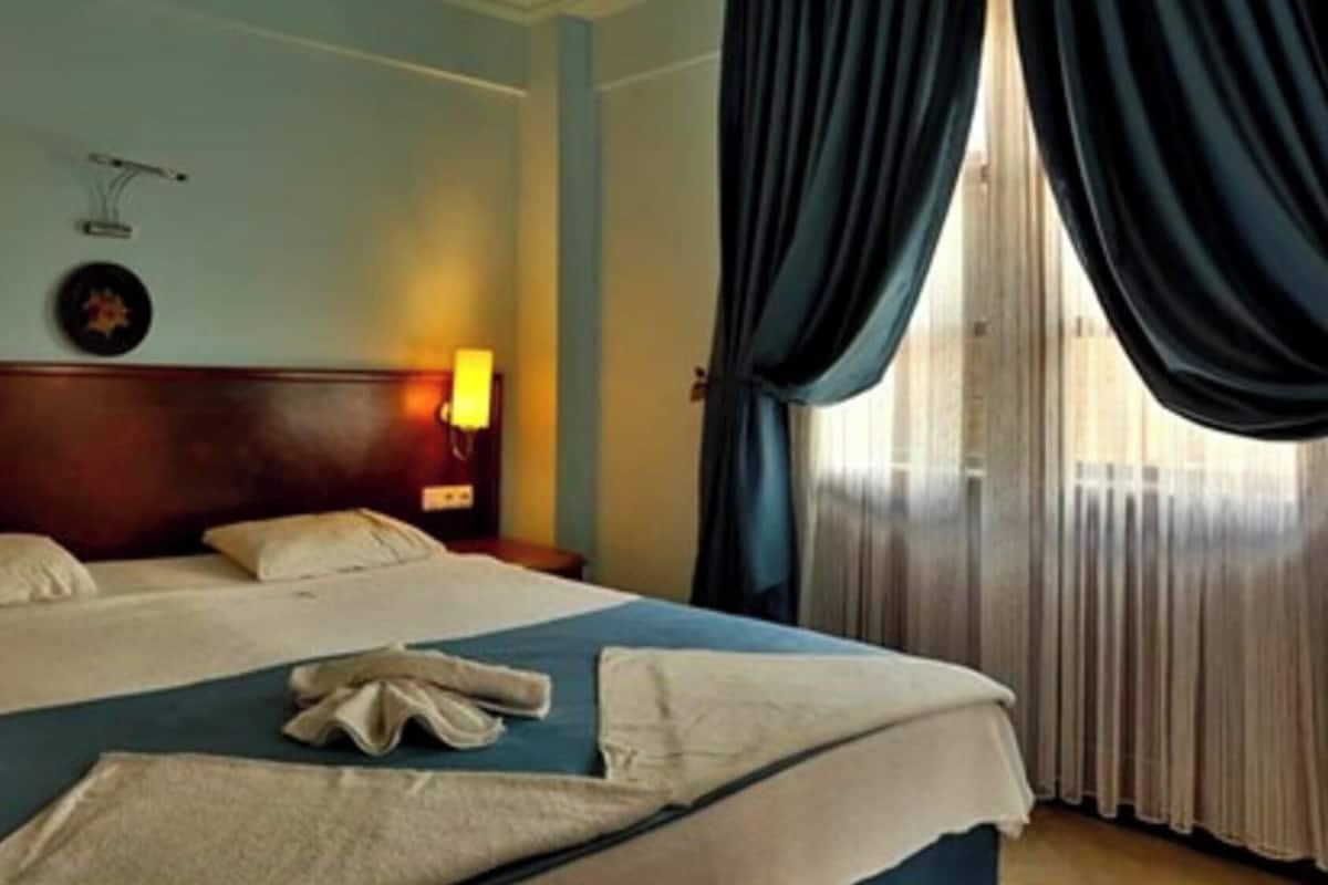 Ein Hotelzimmer mit einem großen Bett, einer Nachttischlampe, einer Wanduhr und einem Fenster mit Vorhängen.