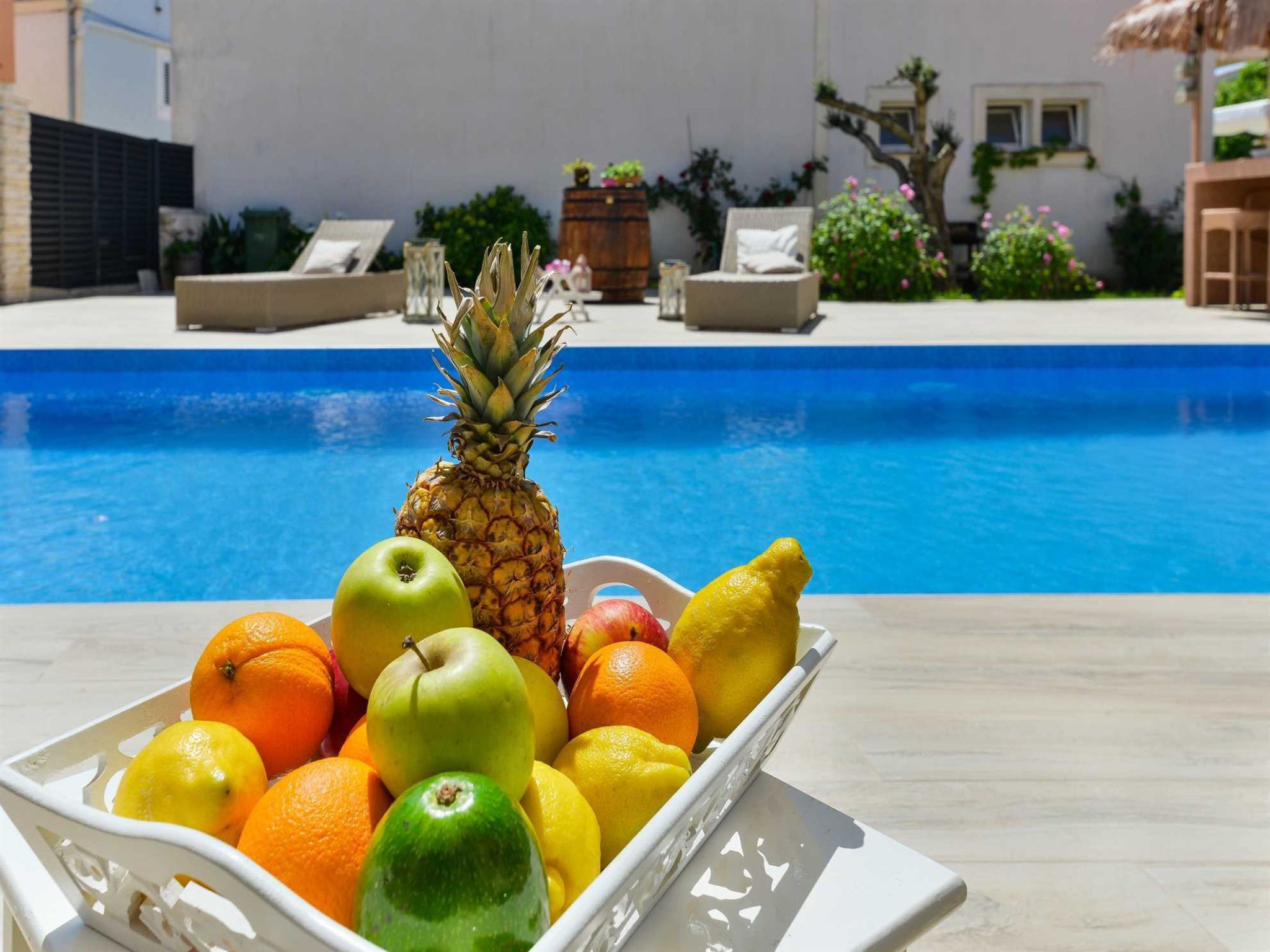 En frugtfat med ananas, æbler, appelsiner og citroner ved en pool.