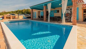 Ein Schwimmbecken mit Fliesenrand, eine überdachte Terrasse mit Sitzgelegenheiten und ein Ziegelgebäude mit blauen Fensterläden.