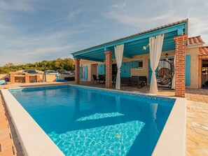 Ein Schwimmbecken mit Fliesenrand, eine überdachte Terrasse mit Sitzgelegenheiten und ein Ziegelgebäude mit blauen Fensterläden.