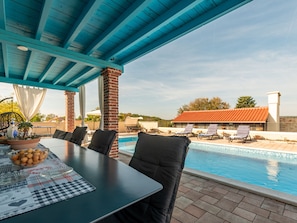 Un espace piscine extérieur doté d’un patio couvert, de chaises longues et d’une table à manger avec un set de table.