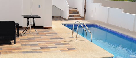 Una piscina con borde de azulejos, una sombrilla blanca y una mesa con sillas.