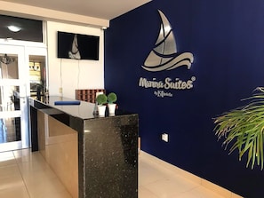 Balcão de recepção com parede azul escuro exibindo o logotipo Marina Suites e um gráfico de um veleiro.