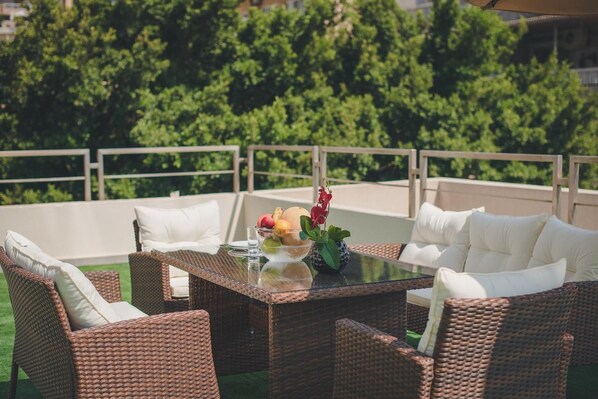A wicker outdoor seating area with a glass table, white cushions, and a fruit bowl. - Louis V Hotel (Dbayeh)