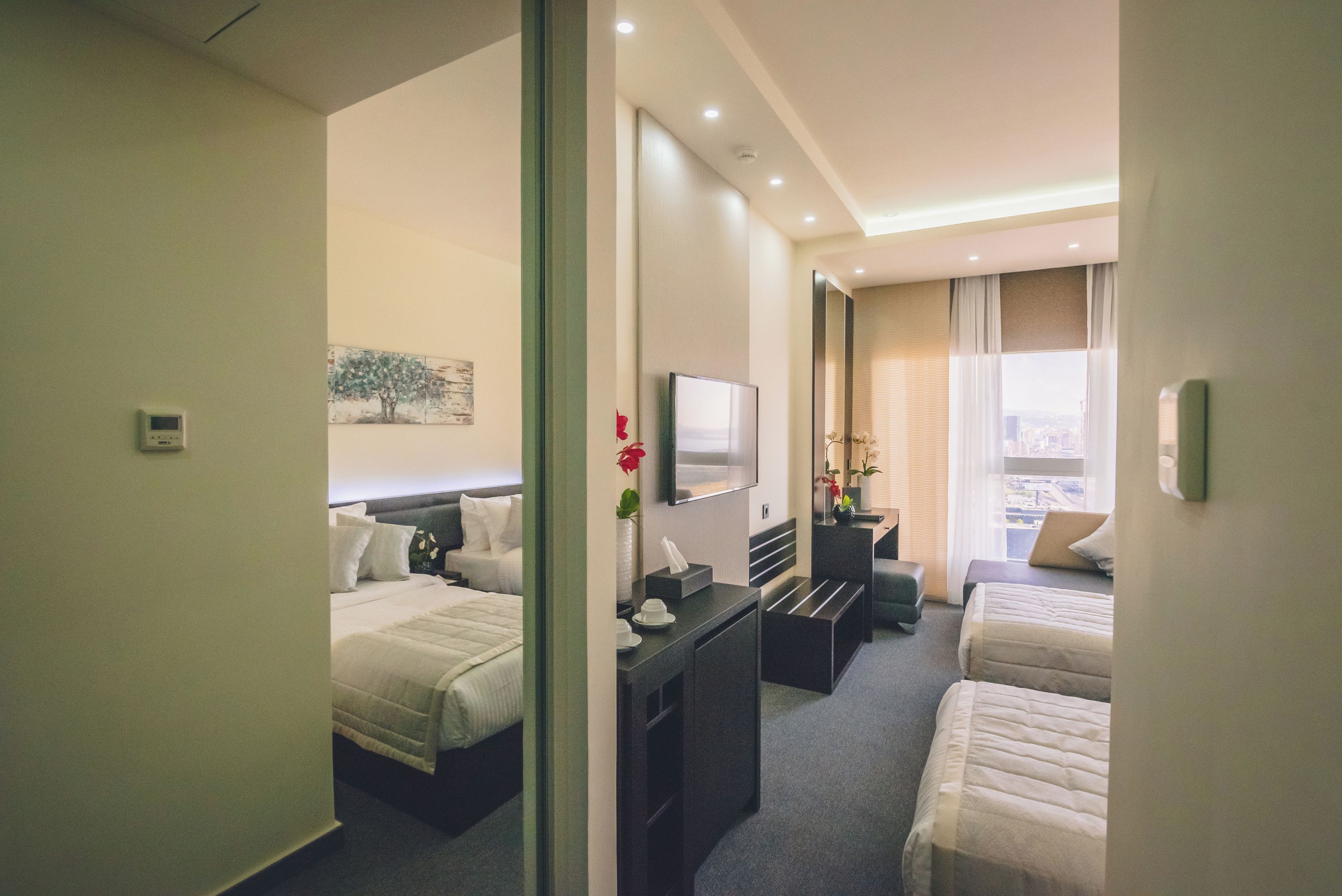 A hotel room with two beds, a desk, a flat-screen TV, a mirror, and a window with a city view.
