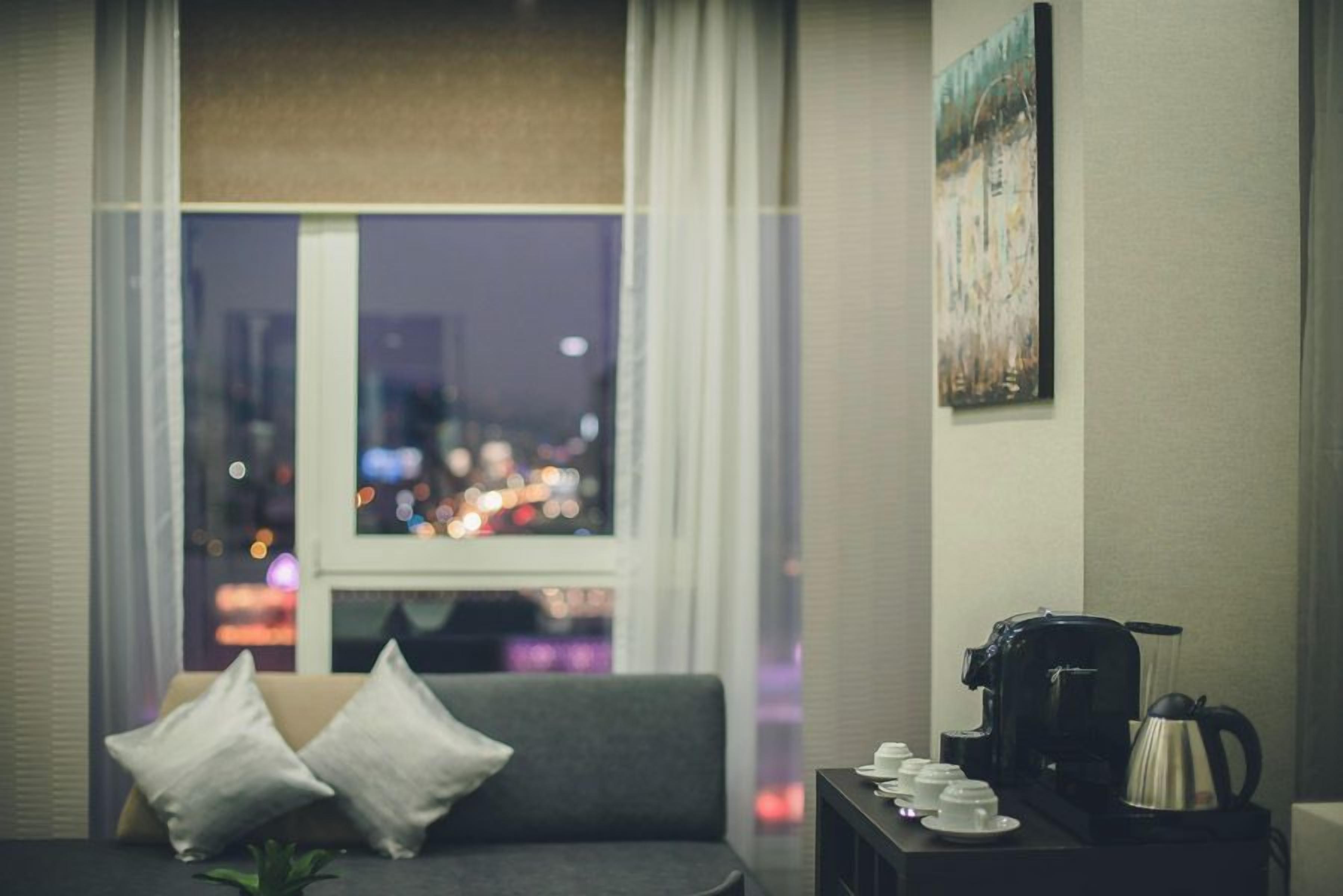 A hotel room with a sofa, pillows, a coffee maker, and a view of a cityscape at night.