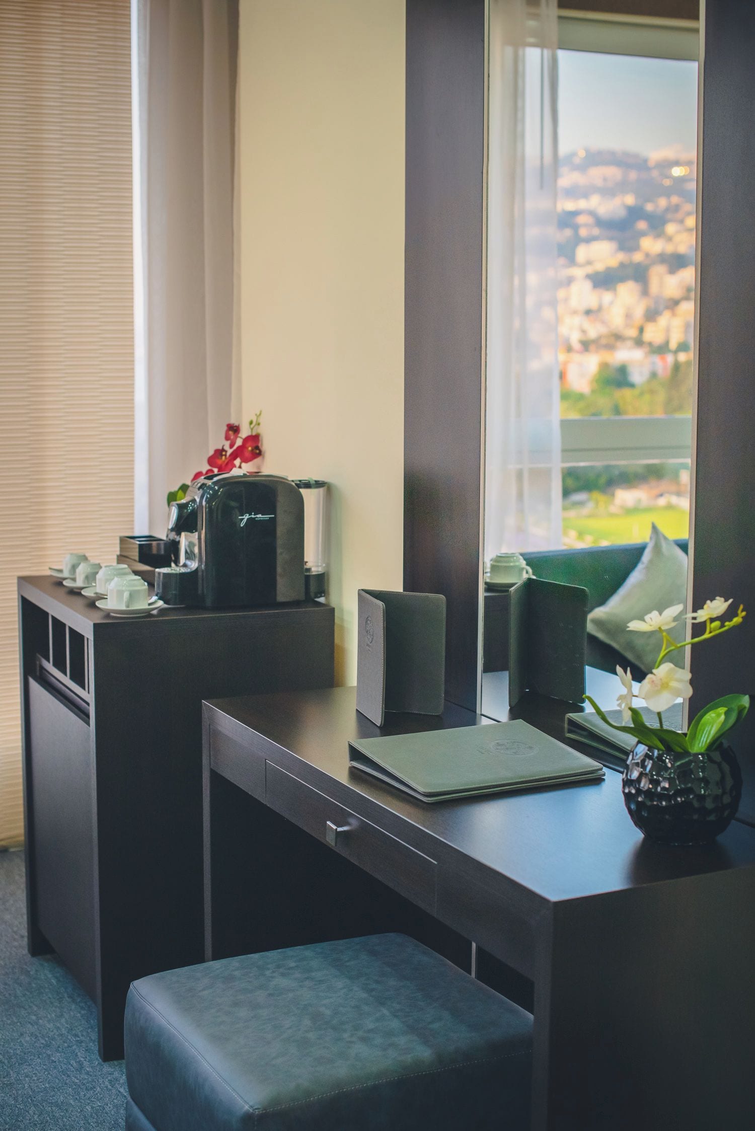 Habitación de hotel con escritorio, cafetera, un jarrón con flores y vista a un paisaje urbano a través de un ventanal grande.