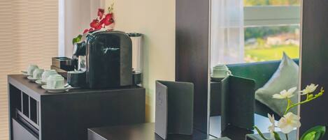 A hotel room with a desk, a coffee maker, a vase with flowers, and a view of a cityscape through a large window.