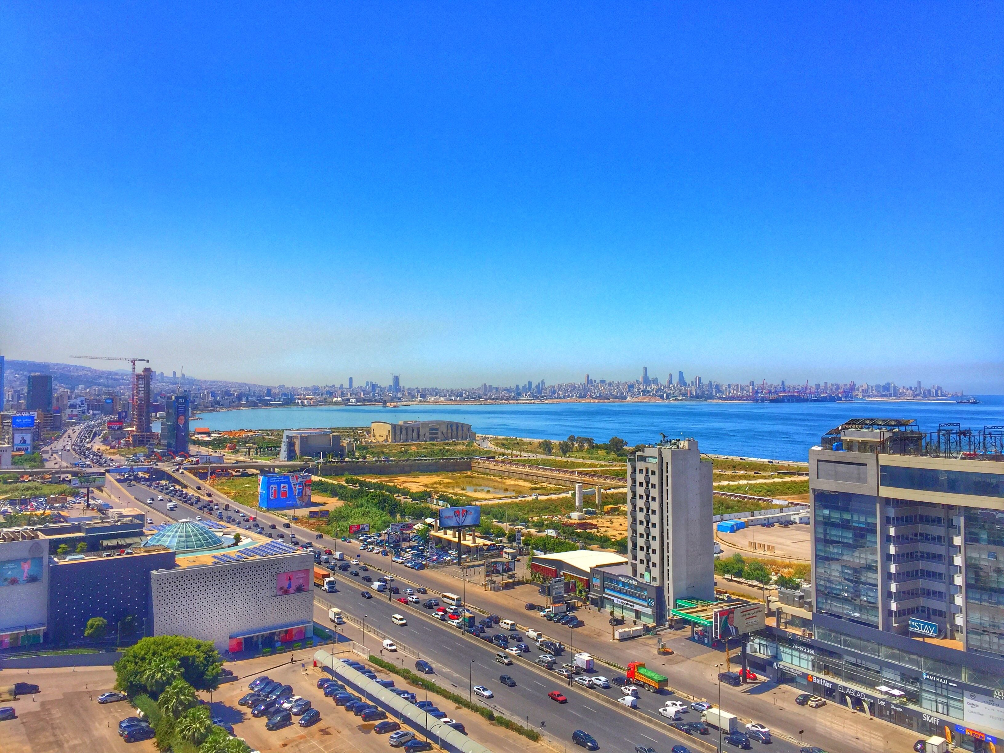 A cityscape with a prominent building featuring a large billboard, a busy road with vehicles, and a clear view of the coastline.