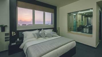 A modern hotel room with a large bed, a nightstand, a window with a city view, and a mirror reflecting the room.