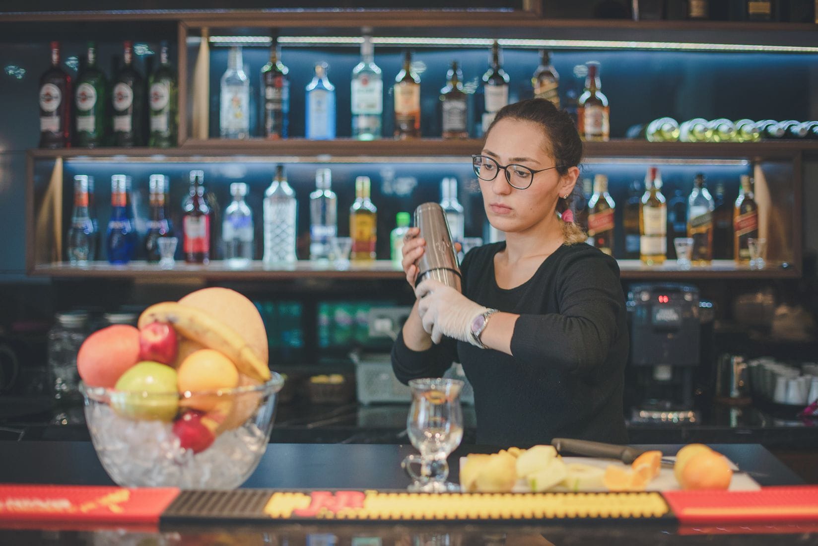 Un barman prépare un cocktail dans un bar, avec une variété de bouteilles en arrière-plan.