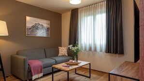A modern living room with a grey sofa, a wooden coffee table, a painting of mountains, and a large window with curtains. - Pradas Resort Brigels, (Breil/Brigels), 65055B by Interhome (Breil/Brigels)