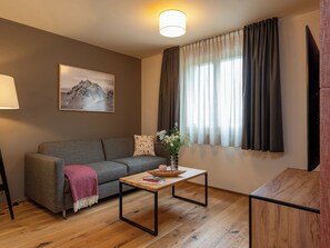Ein modernes Wohnzimmer mit grauem Sofa, einem Holztisch, einem Bergpanorama und einem großen Fenster mit Vorhängen.