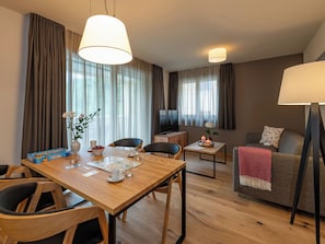 A modern hotel room with a wooden dining table, chairs, a sofa, a TV, and a lamp.
