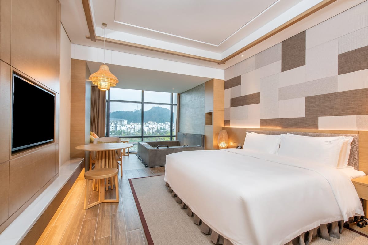 Una habitación de hotel moderna con una cama grande, un televisor montado en la pared, un área de comedor con mesa y sillas, y vistas a la ciudad a través de amplios ventanales.