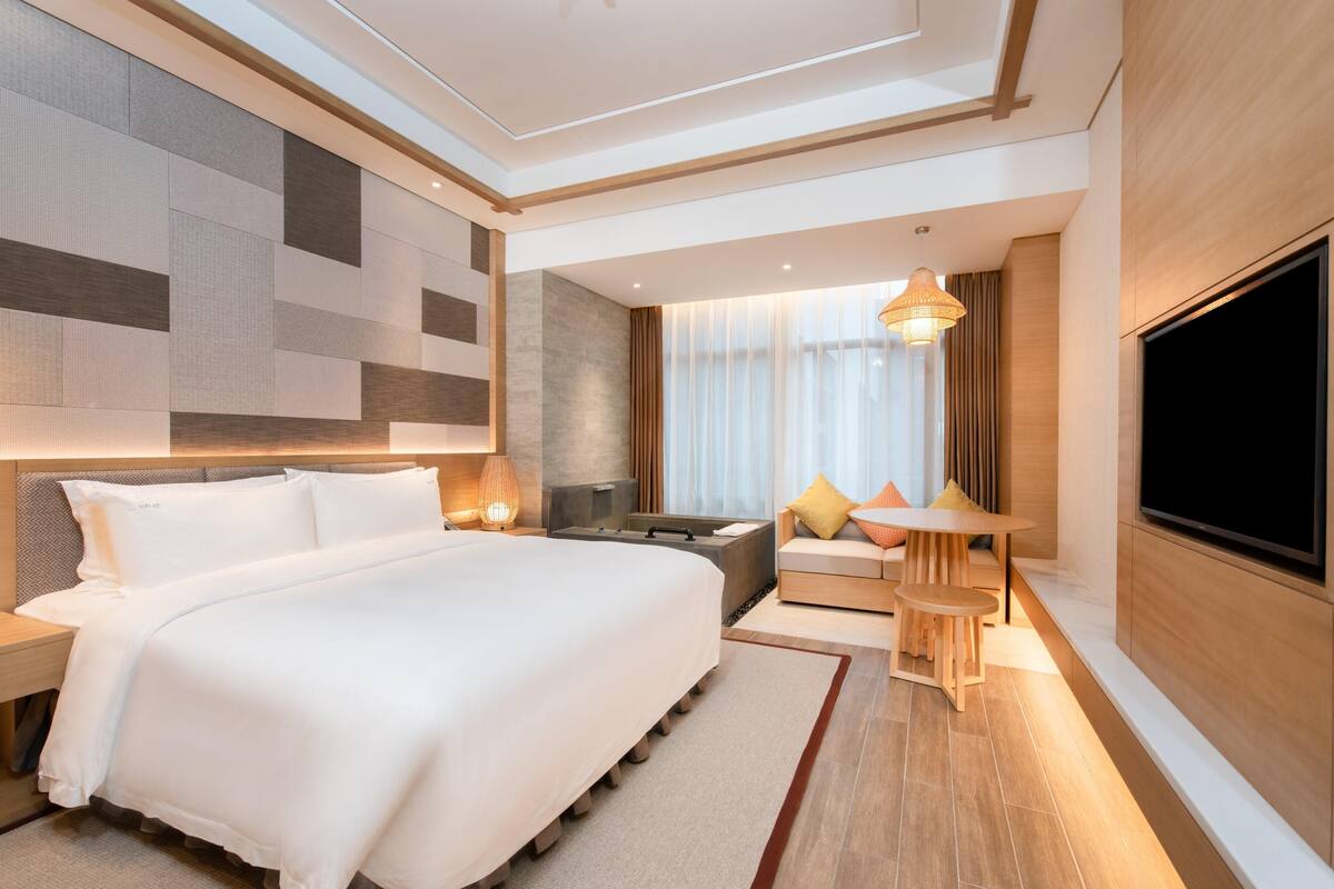 Una habitación de hotel moderna con una cama grande, una zona de estar con una mesa, un televisor y un ventanal con cortinas.