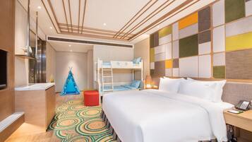 A hotel room with a bunk bed, a desk with a lamp, a small red stool, and a colorful rug.