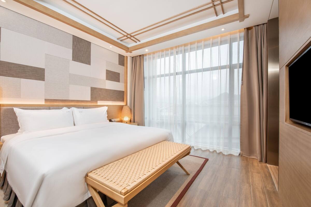 Una habitación de hotel moderna con una cama grande, un banco, un televisor y un ventanal con cortinas.