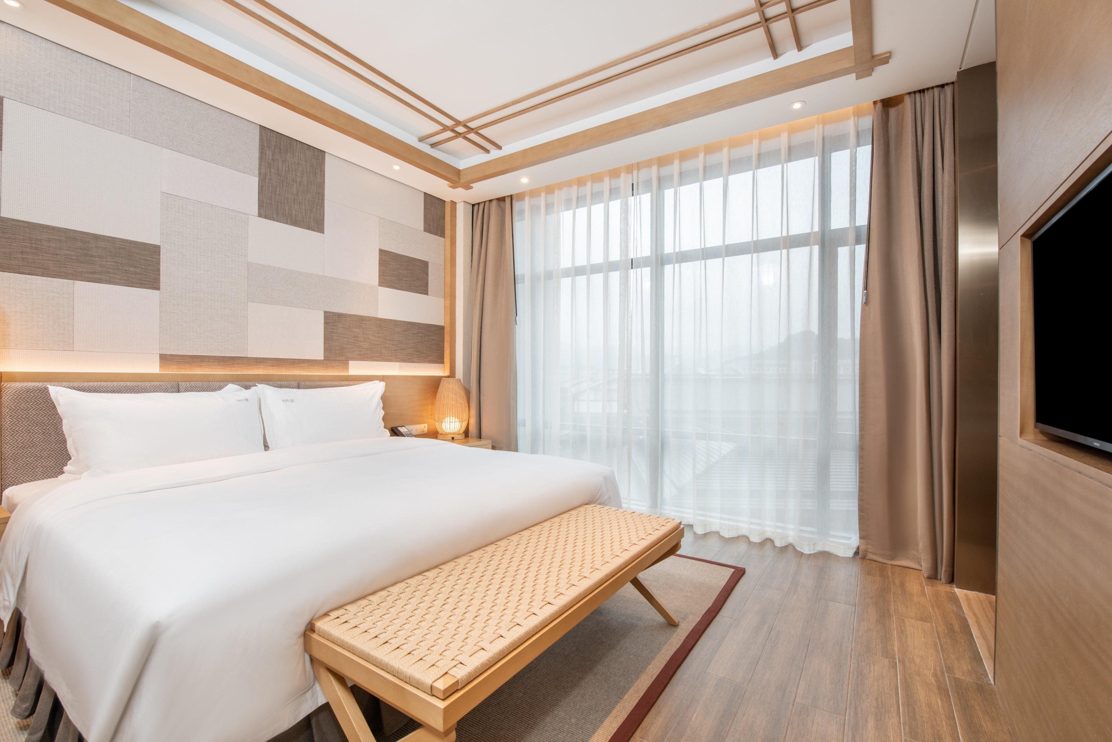 Una habitación de hotel moderna con una cama grande, un banco, un televisor y un ventanal con cortinas.