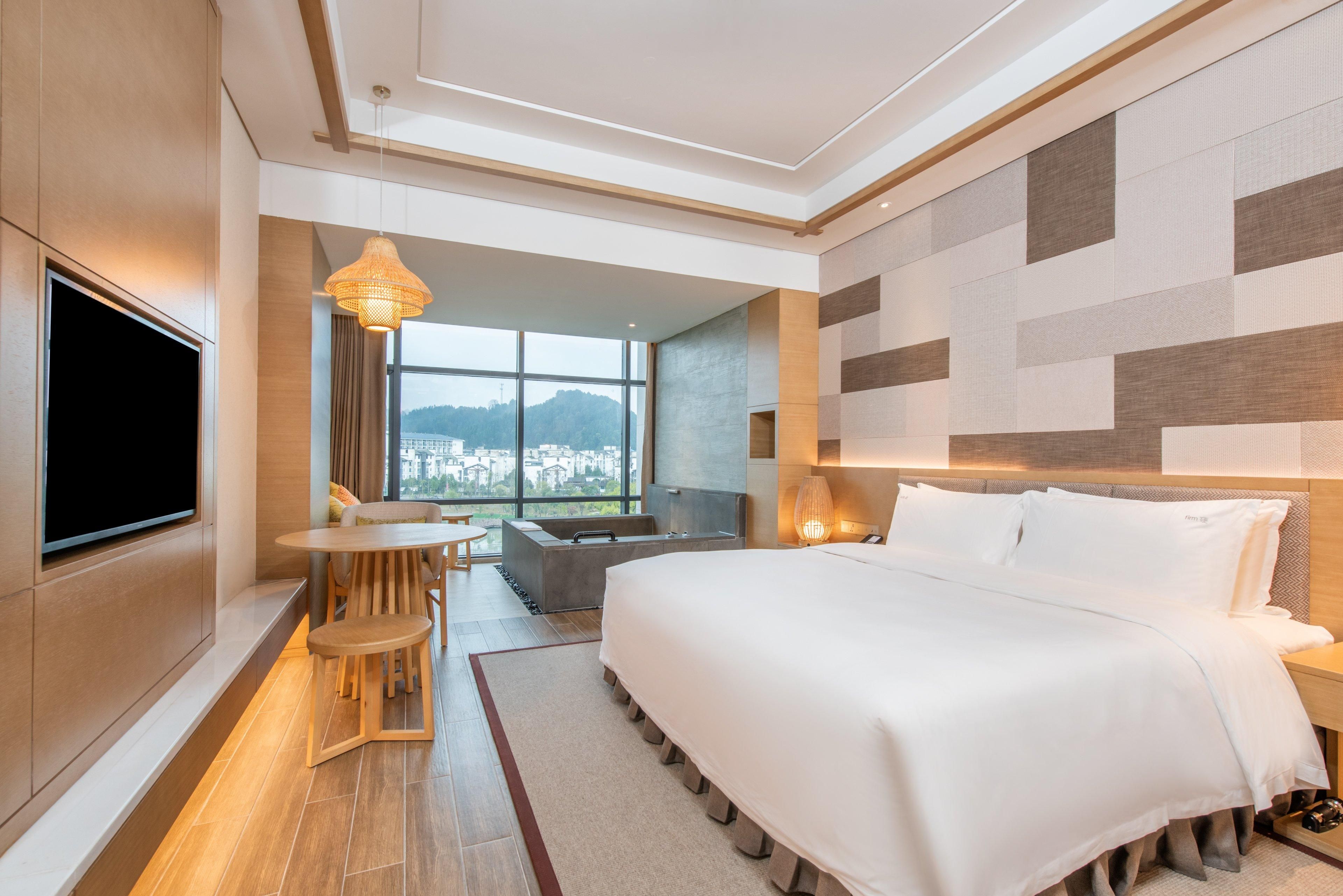 Una habitación de hotel moderna con una cama grande, un televisor montado en la pared, un área de comedor con mesa y sillas, y vistas a la ciudad a través de amplios ventanales.