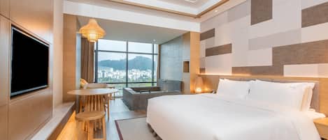 A modern hotel room with a large bed, a TV mounted on the wall, a dining area with a table and chairs, and a view of the city through large windows.