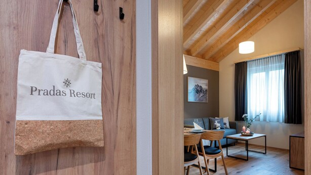 Un intérieur en bois comprenant un coin repas, un canapé et une fenêtre avec des rideaux. Un sac fourre-tout blanc orné de l’inscription « Pradas Resort » est accroché au mur.
