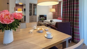 Habitación de hotel con una mesa de comedor de madera, un ventanal con cortinas a cuadros, una cama con televisor y un jarrón con flores.