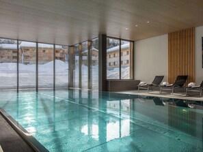 An indoor swimming pool with clear glass walls, lounge chairs, and a view of buildings outside. - Pradas Resort Brigels, (Breil/Brigels), 65055B by Interhome (Breil/Brigels)