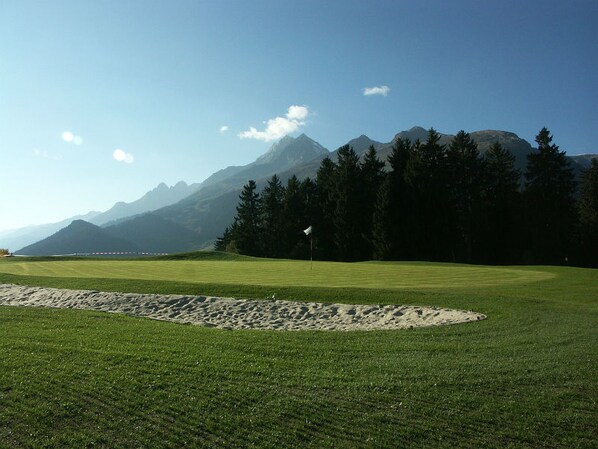 Um campo de golfe com uma bandeira, um bunker de areia e montanhas ao fundo.
